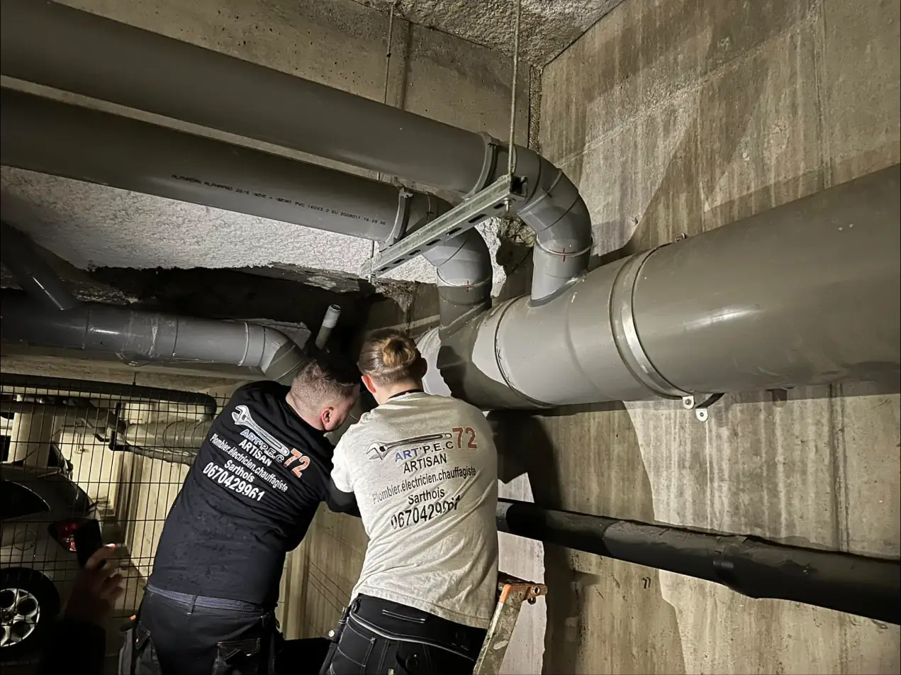 Deux artisans d'Art'Pec 72, en t-shirts à leurs couleurs, sur un réseau d'évacuation en PVC dans un parking souterrain